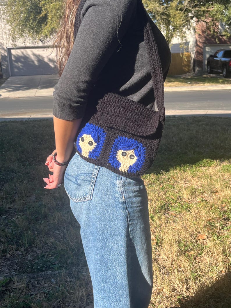 May include: A black crocheted shoulder bag with two blue and white embroidered faces. The bag has a long strap.