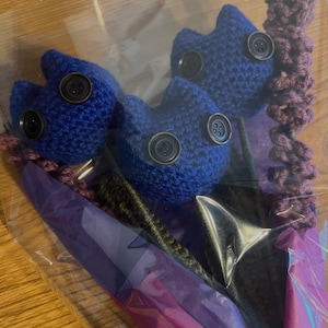 May include: A bouquet of three blue crocheted flowers with black buttons for eyes. The flowers are wrapped in purple and green tissue paper and placed in a clear plastic bag.