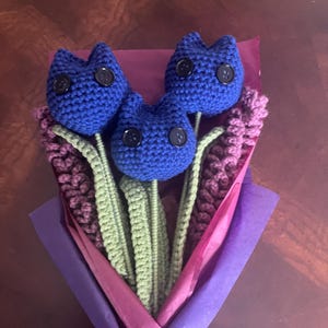May include: A bouquet of three blue crocheted tulips with black button eyes, wrapped in purple and pink tissue paper.