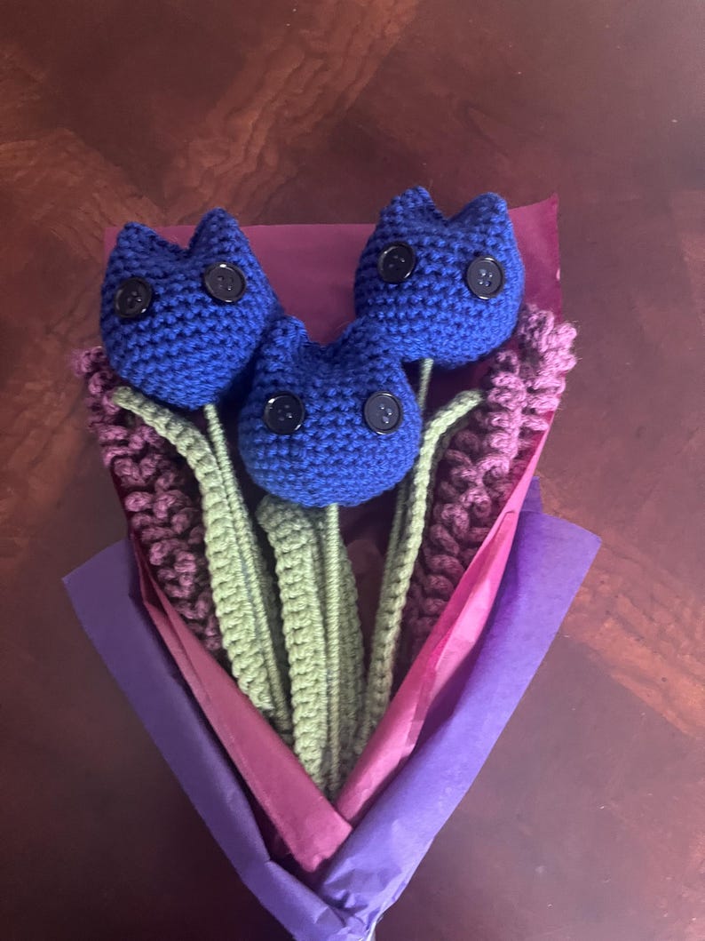 May include: A bouquet of three blue crocheted tulips with black button eyes, wrapped in purple and pink tissue paper.