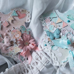 May include: Two handmade baby shower decorations shaped like onesies. The onesie on the left is pink with a bear, butterflies, and flowers. The onesie on the right is blue with a bunny, a bird, and flowers.