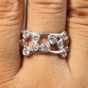 May include: A silver-toned band ring featuring an openwork design with two parallel horizontal bars connected by multiple circular settings, each containing a single clear round-cut stone. The metal has a polished finish and a wide profile.
