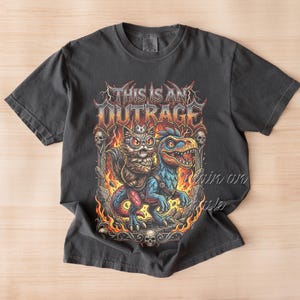 May include: Dark gray t-shirt featuring a colorful graphic of a cat wearing goggles riding a blue dinosaur, with the text "THIS IS AN OUTRAGE" in a fiery font. The design includes skulls and flames, creating a bold, eye-catching design.