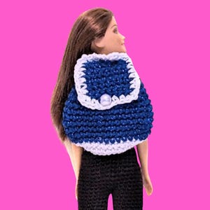 May include: A doll with long brown hair wearing a dark blue crocheted backpack with a white trim and a single white pearl-like button closure. The doll is also wearing black crocheted trousers. The backpack is positioned on the back of the doll.
