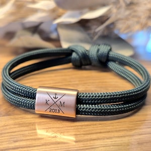 Könnte beinhalten: Dunkelgrünes Seilarmband mit kupferfarbener Schließe. Die Schließe zeigt ein Ankersymbol, die gekreuzten Buchstaben "K" und "M" und das Jahr "2013". Das Armband ist oben geknotet.