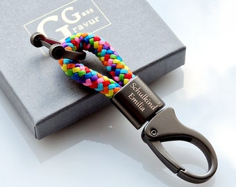 May include: A keychain featuring a braided cord in vibrant rainbow colors attached to a dark metallic clasp. The metal component is engraved with the text Schulkind Emilia. The design includes a dark metal loop and a spring-loaded hook mechanism.
