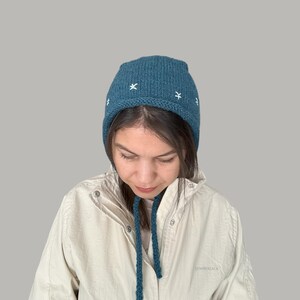 May include: A teal blue knitted hat with a white embroidered star pattern. The hat has a long tie that can be tied under the chin.