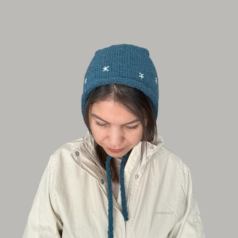 May include: A teal blue knitted hat with a white embroidered star pattern. The hat has a long tie that can be tied under the chin.