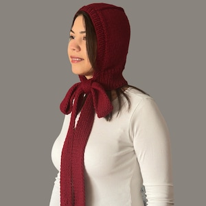 May include: A knitted claret hood with long ties. The hood covers the head and neck, with a tie closure at the front. The knitted fabric has a textured appearance. The hood is a winter accessory.