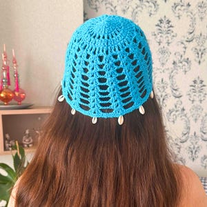 May include: A blue crocheted hat with white shell accents. The hat is worn on a person with long brown hair.