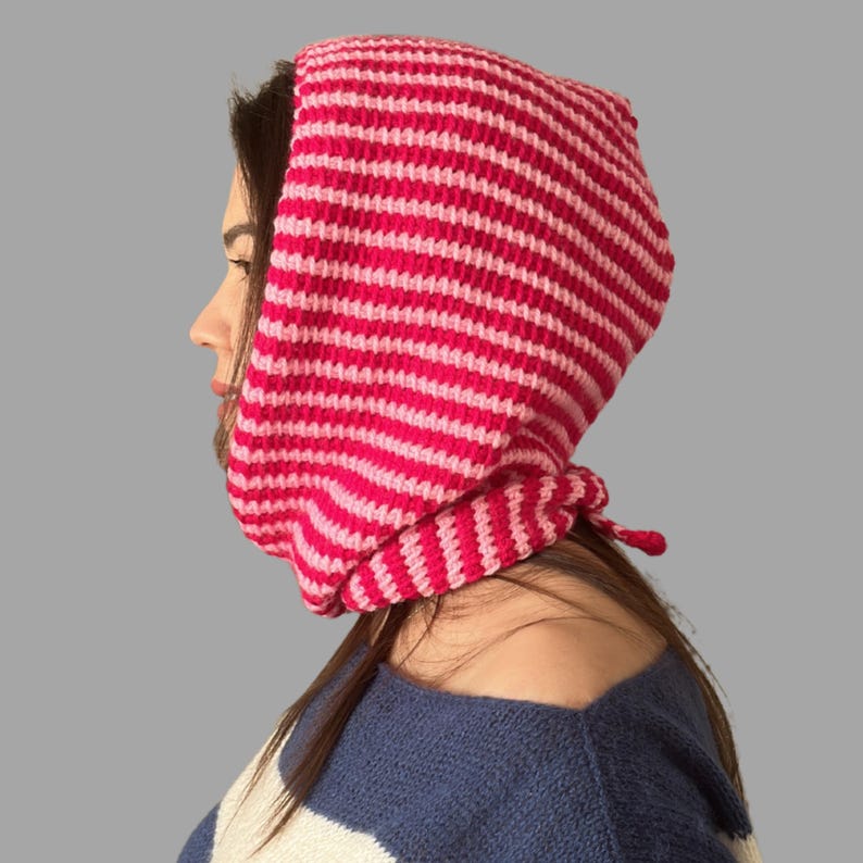 May include: A pink and light pink striped crochet bonnet. The bonnet is tied at the neck and covers the head and neck. The person in the image is wearing a blue and white striped sweater.
