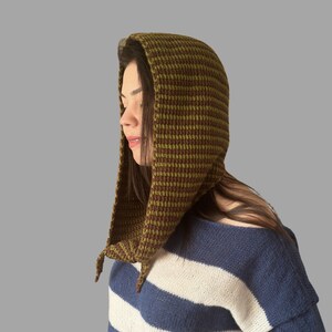 May include: A knitted hood in shades of olive green and brown, with a textured pattern. The hood is worn over the head and drapes down the shoulders. The person is wearing a blue and white striped sweater.