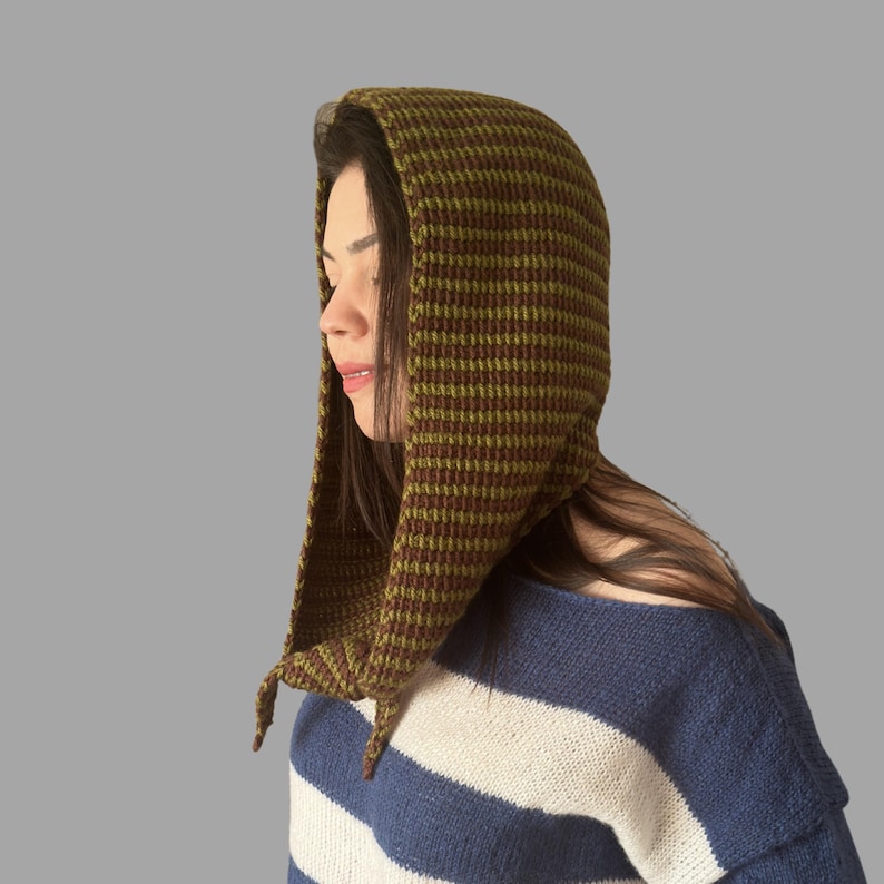 May include: A knitted hood in shades of olive green and brown, with a textured pattern. The hood is worn over the head and drapes down the shoulders. The person is wearing a blue and white striped sweater.