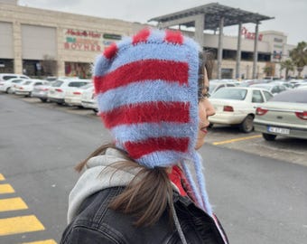 May include: A blue and red striped knitted hat with a fluffy texture. The hat has a long tie that hangs down the back.