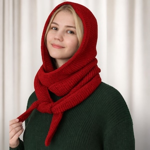 Könnte beinhalten: Ein gestricktes rotes Kapuzen-Schal-Set. Die Kapuze ist hochgezogen und rahmt das Gesicht ein, und der Schal umschlingt den Hals mit langen Bändern. Das Strickwerk hat eine kräftige, einfarbige rote Farbe, und die Textur des Strickens ist sichtbar.