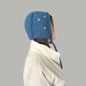 May include: A blue knitted hat with white embroidered stars. The hat has a long tie that hangs down the back of the neck.