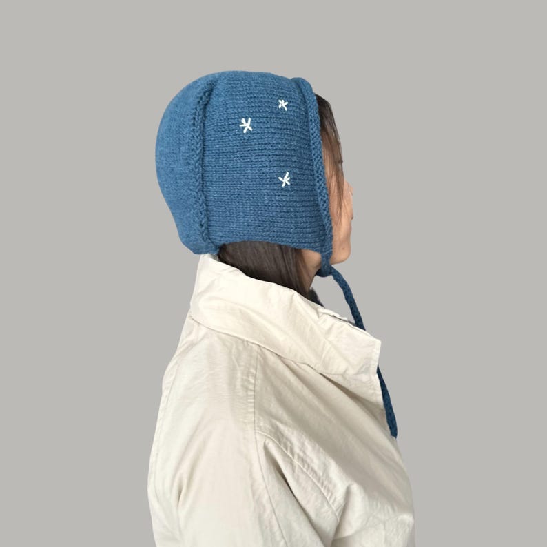 May include: A blue knitted hat with white embroidered stars. The hat has a long tie that hangs down the back of the neck.