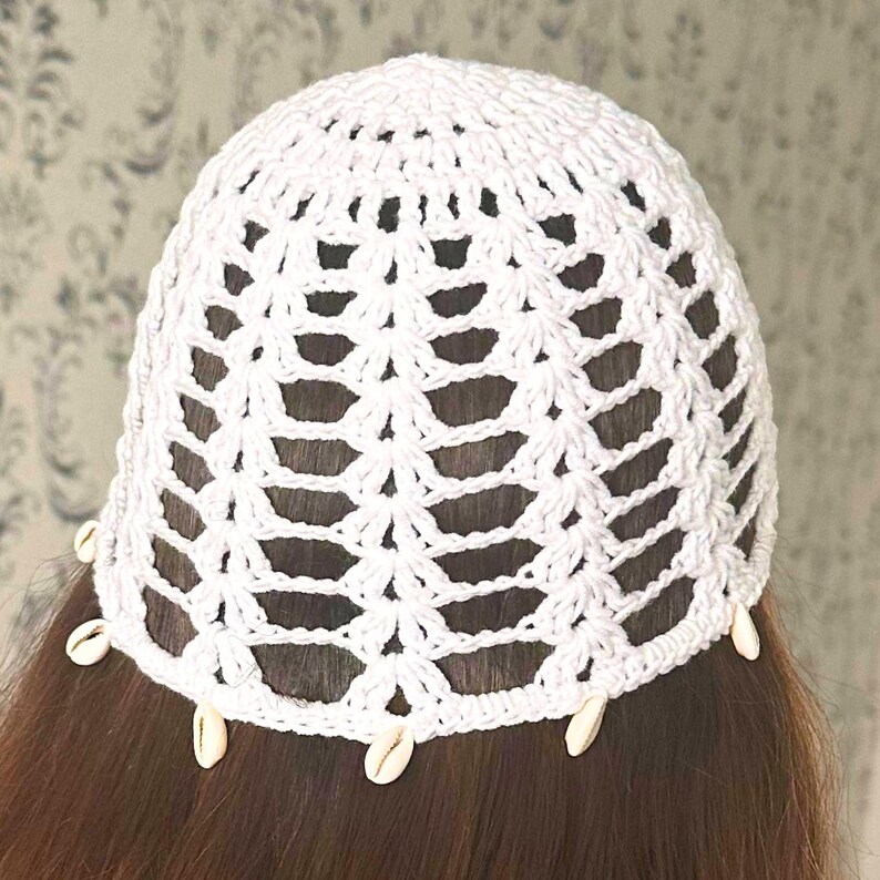 May include: White crochet beanie hat with a delicate openwork design and white seashells attached around the brim.