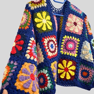 May include: A handmade, open-front crochet cardigan in a vibrant patchwork design. The base color is a deep blue, with colorful floral squares in shades of yellow, pink, orange, purple, and red. The cardigan is displayed on a blue hanger.