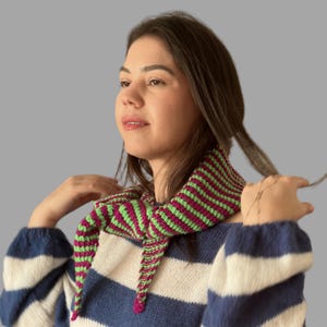 May include: A striped crochet scarf in shades of green and magenta. The scarf is worn around the neck and has a triangular shape. The person in the image is wearing a blue and white striped sweater.