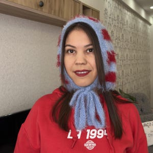 May include: A woman wearing a blue and red striped knit hat with a matching scarf. The hat has a fluffy texture and covers her ears. She is wearing a red hoodie with the text "L7 1994" and a small globe graphic.