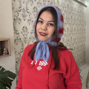 May include: A woman wearing a red hoodie with the text "27 199" and a globe graphic. She is wearing a blue and red striped knit hat with a fluffy texture.