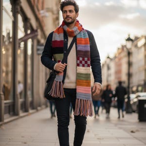 Może przedstawiać: Wielokolorowa, pasiasta szalik z frędzlami jest noszony wokół szyi. Szalik ma różne kolory, w tym pomarańczowy, szary, czerwony i fioletowy. Osoba nosi dżinsową kurtkę, kremowy sweter i czarne spodnie. Czarna torba kurierska jest przewieszona przez ramię.