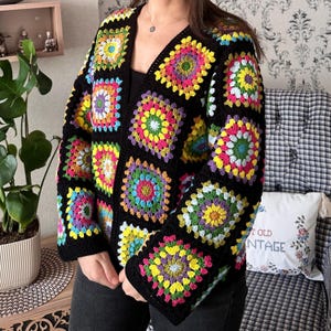 Puede incluir: Una chaqueta de punto de ganchillo negra con motivos de cuadrados de la abuela coloridos. La chaqueta tiene cuello en V y mangas largas.