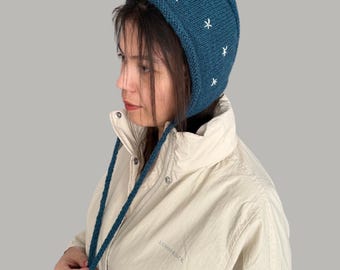 May include: A teal blue knitted hat with white snowflake embroidery. The hat has a long tie that can be tied under the chin.