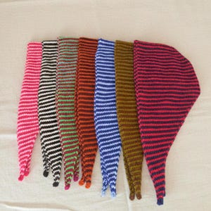 May include: A collection of seven knitted scarves, each with a unique color scheme of horizontal stripes. Colors include pink, black and white, green and red, orange, blue and white, brown and navy, and red and navy. The scarves are arranged side by side.