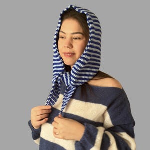 May include: A blue and white striped knit hood with ties, worn over a matching striped sweater. The hood is made of a soft, textured material and is designed to cover the head and neck.