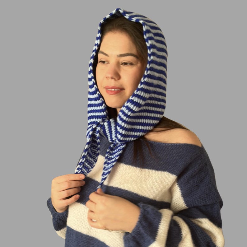 May include: A blue and white striped knit hood with ties, worn over a matching striped sweater. The hood is made of a soft, textured material and is designed to cover the head and neck.