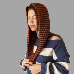 May include: An orange and brown striped knit hood. The hood is worn over the head and drapes down the front. The person in the image is wearing a blue and white striped sweater. The hood is a warm accessory.