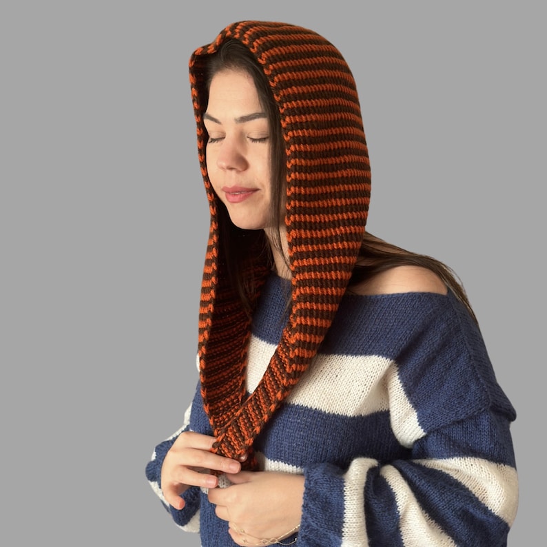 May include: An orange and brown striped knit hood. The hood is worn over the head and drapes down the front. The person in the image is wearing a blue and white striped sweater. The hood is a warm accessory.