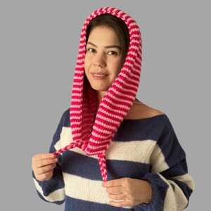 May include: A pink and white striped knitted hood with drawstrings. The hood is worn over a blue and white striped sweater. The hood is made of a soft, textured yarn.
