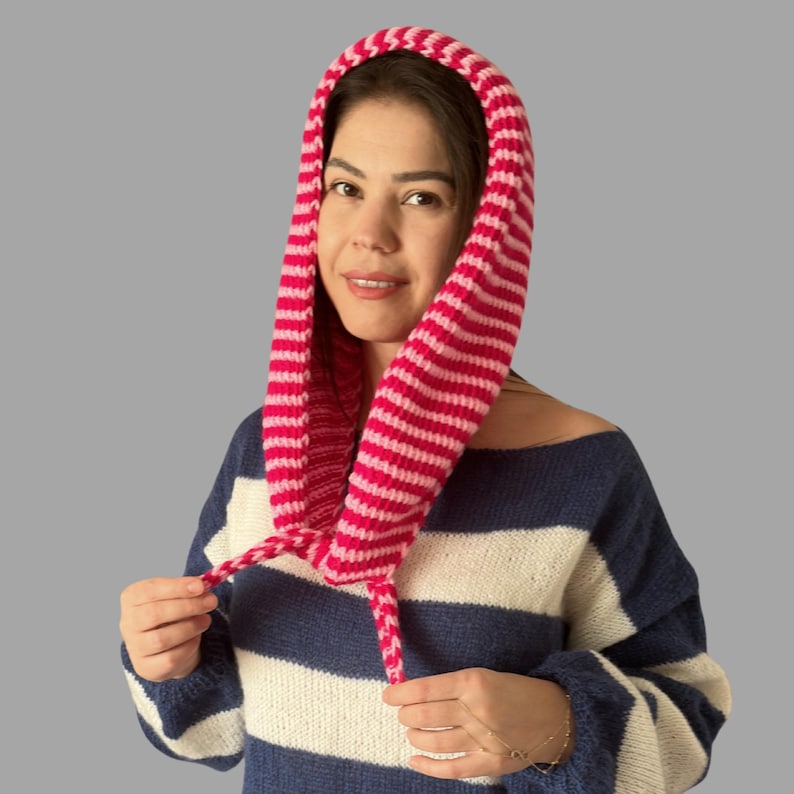 May include: A pink and white striped knitted hood with drawstrings. The hood is worn over a blue and white striped sweater. The hood is made of a soft, textured yarn.