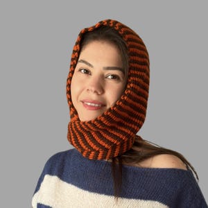 May include: A knitted hood in brown and orange stripes. The hood is worn over the head and around the neck. The person in the image is wearing a blue and white striped sweater.