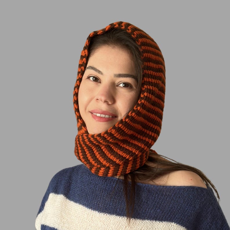 May include: A knitted hood in brown and orange stripes. The hood is worn over the head and around the neck. The person in the image is wearing a blue and white striped sweater.