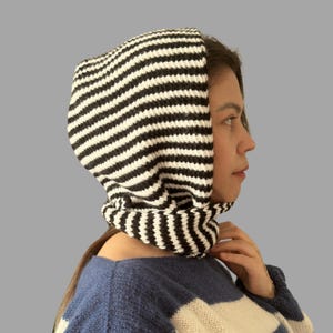 May include: A black and white striped knit hood and cowl. The hood is pulled up, covering the head and neck, with the cowl resting around the neck. The knitwear features horizontal stripes. The person is wearing a blue and white striped sweater.
