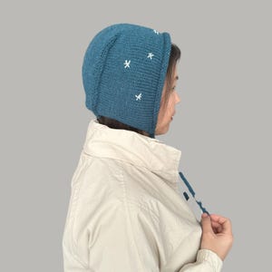May include: A teal blue knitted hat with white embroidered stars. The hat has a long flap that can be tied under the chin.