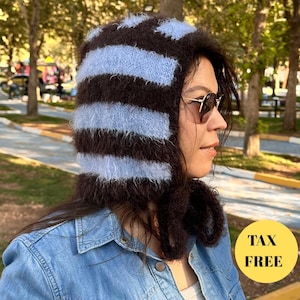 May include: A cozy, striped knit hat in brown and light blue. The hat is worn by a person wearing sunglasses and a denim jacket. The hat has a hood and a scarf-like extension. The image includes a yellow circle with the text "TAX FREE".