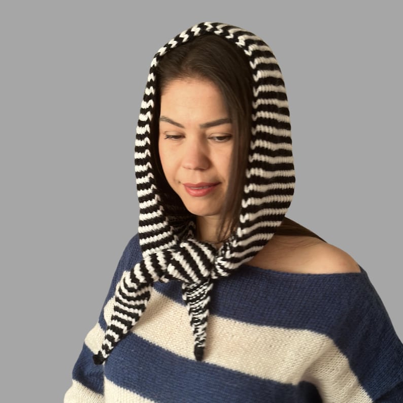 May include: A black and white striped knitted hood and scarf combination. The hood is pulled up, and the scarf is tied at the neck. The person is wearing a blue and white striped sweater. The hood and scarf are made of a soft, textured yarn.