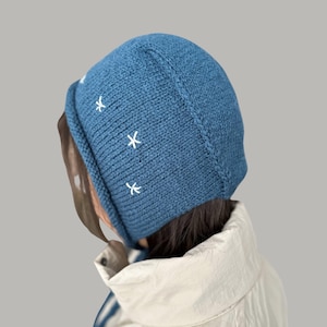 May include: A blue knitted beanie with white embroidered stars. The beanie is folded down and has a loose fit.