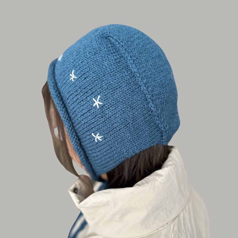 May include: A blue knitted beanie with white embroidered stars. The beanie is folded down and has a loose fit.