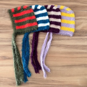 May include: Four colorful, striped knit hats with long, dangling ties. The hats feature green with red stripes, blue with white stripes, purple with white stripes, and yellow with purple stripes. The hats are displayed on a light brown wooden surface.