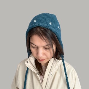 May include: A teal blue knitted hat with white embroidered stars. The hat has a long, pointed flap that can be tied under the chin. The hat is worn by a person with long brown hair.