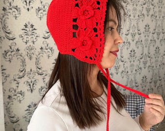 May include: A red crocheted bonnet with a flower design and ties. The bonnet is worn by a person with long brown hair.