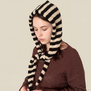 May include: A black and cream striped knitted hood with long ties. The hood is made of a soft, textured material and is designed to be worn over the head and around the neck. The stripes are evenly spaced, creating a bold pattern.