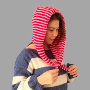 May include: A pink and white striped knitted hood. The hood is pulled over the head and tied at the neck. The person in the image is wearing a blue and white striped sweater.