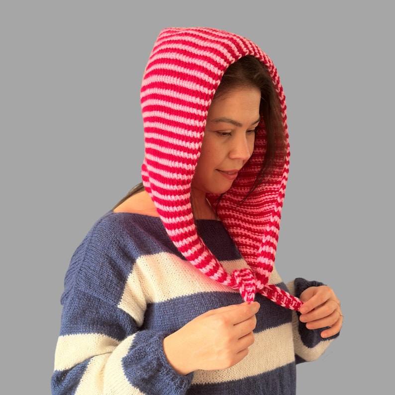 May include: A pink and white striped knitted hood. The hood is pulled over the head and tied at the neck. The person in the image is wearing a blue and white striped sweater.
